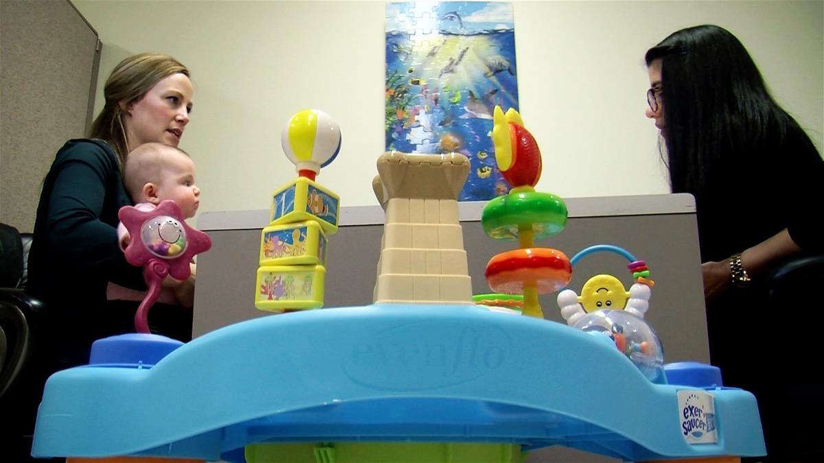 A mom holds a baby while talking to an IKIDS researcher. A baby toy sits in the foreground.